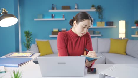 Mujer-Joven-Asiática-Que-Trabaja-Desde-Casa-Tomando-Notas-Cuidadosamente-Y-Trabajando-Desde-Una-Computadora-Portátil.
